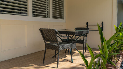 Wicker plastic furniture stands on the outdoor terrace against the wall - a table, two chairs. Blinds on the windows. Green vegetation is nearby. Philippines.
