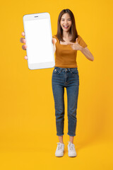 Beautiful Asian woman pointing finger on mobile and holding big smartphone mockup of blank screen on yellow background.
