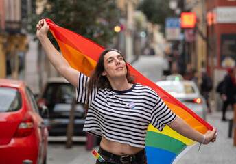 A transgender woman holding a rainbow flag in a city street. The flag is a symbol of the LGBTQ+ community