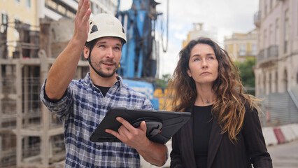 Engineer constructor discuss build plan with investor woman. Builder guy show construction site project tablet pad. Worker tell blueprint client. Man work city street Customer look at new house scheme