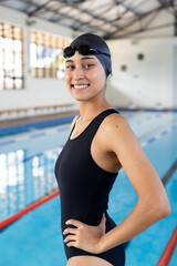 Biracial young female swimmer standing by pool indoors, wearing black swimsuit