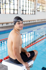 Caucasian young male swimmer sitting by pool indoors, looking at camera, smiling