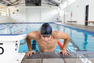 Caucasian young male swimmer resting at swimming pool edge indoors, wearing goggles