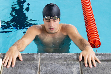 Caucasian young male swimmer resting at swimming pool edge indoors, looking focused