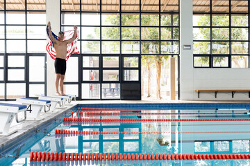 Caucasian young male swimmer stretching on pool edge indoors, holding american flag, copy space