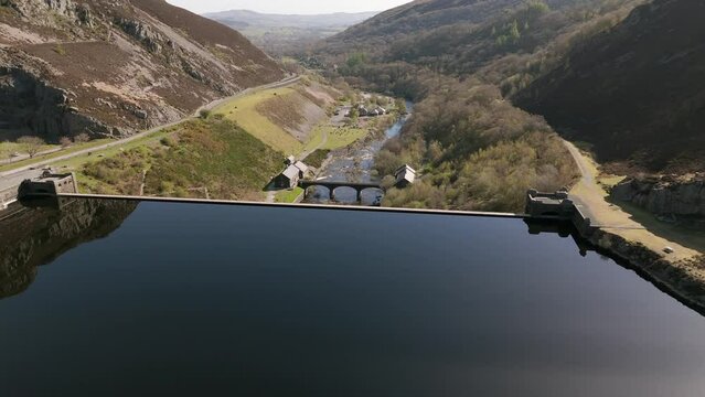 An aerial view of Caban Coch dam and reservoir on a sunny spring day in the Elan valley, Powys, Wales. Flying over the reservoir towards the dam with the visitors centre in the distance