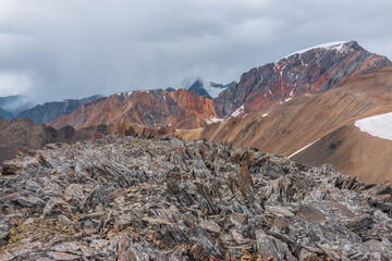 Awesome top view from rocky pass to sharp rocks on ridge of vivid colors against snowy peak in low clouds. Scenic landscape with colorful sheer crags and snow-capped mountain top under gray rainy sky. - 793609101