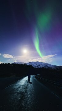 Man gazing up at the aurora dancing around the moon from a road in northern Norway.