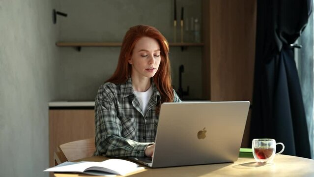 Successful young business woman having cash dollar rain falling while working typing on Apple MacBook laptop sitting at home office desk. Happy businesswoman celebrating success, winning, good news.