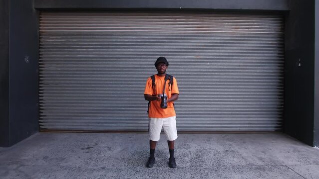 African-American man stands in front of garage roller shutter door holding camera and then stares with direct eye gaze - handheld shot