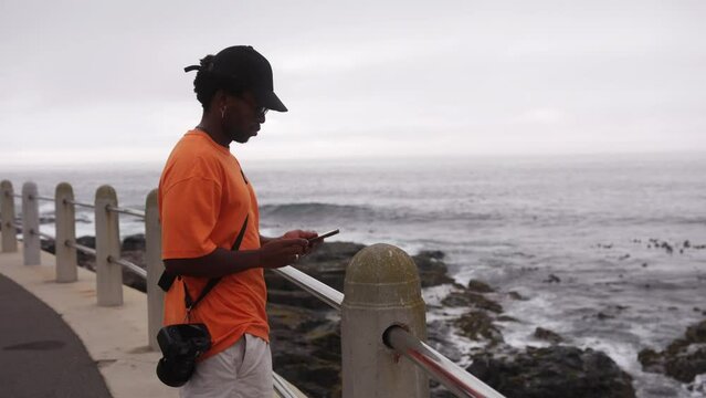 Slow motion shot of young Black man taking phone from pocket to take photos of sea view in Cape Town, South Africa