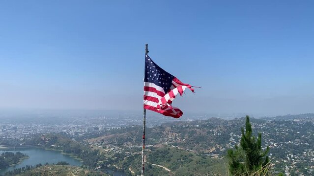 American flag on a hilltop waving, with a lake, mountain city and blue sky in the background