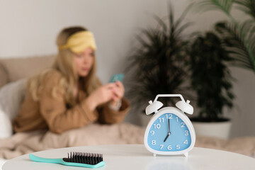 Blurred woman with phone, hairbrush and alarm clock in focus.