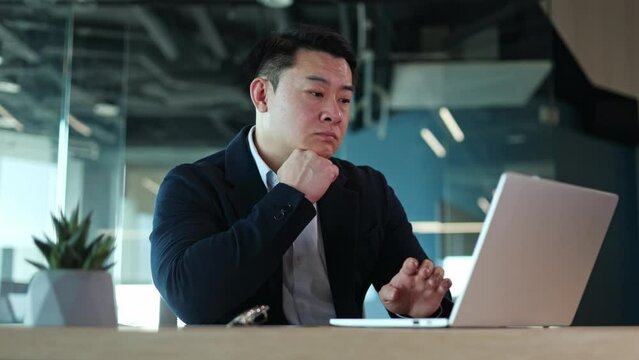Chinese male employee sitting at desk with portable laptop and feeling bored with routine work. Unhappy man in office outfit typing on keyboard with one hand and expressing disappointment with job.