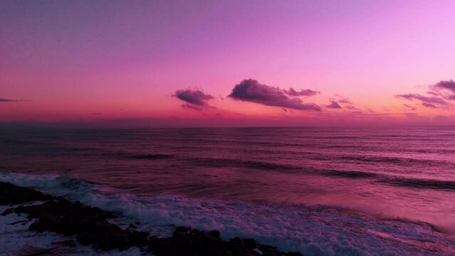 Drone of an epic tranquil scenery of sea waves with beachgoers at sunset time with pink-purple sky