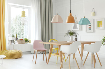 A bright and airy dining room with pastel colored chairs around an oak table, complemented in the style of yellow accents like pendant lights and decor items. The floor is covered in white ceramic til