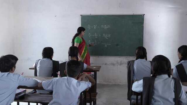 Indian female math teacher writes with chalk on the blackboard teaching sums to elementary students studying at classroom. Education india.