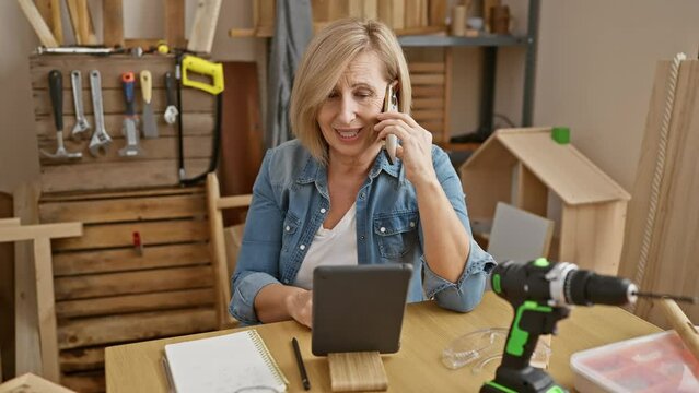 A mature woman in a carpentry workshop communicates on a smartphone while viewing a digital tablet amidst woodworking tools.