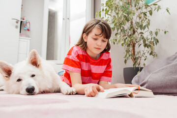 A teenage girl lies on the floor in her room and reads a book, next to her lies a dog, a white...
