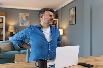 Shot of a businessman suffering from a backache while working at his desk in his office. Man having back pain while working on laptop from home