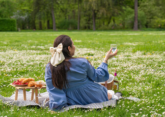 Happy woman enjoys life. Spends time in nature. A lady is sitting on the grass in a park, with a picnic table with food nearby. Back view. There is a white bow on the head. Holding a glass in his hand