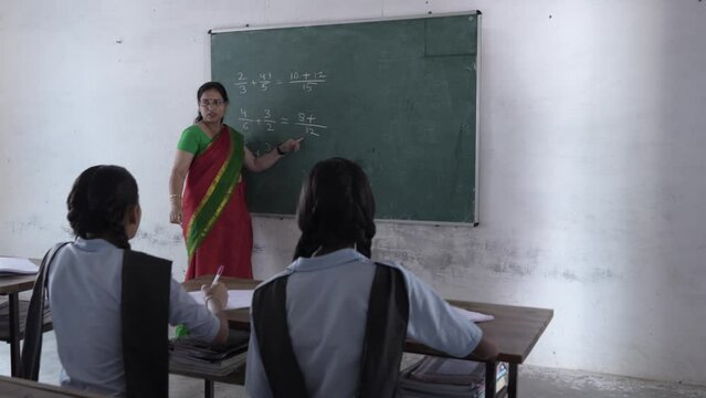Indian female teacher writes with chalk on the blackboard teaching math sums to elementary students studying at classroom. Education india.