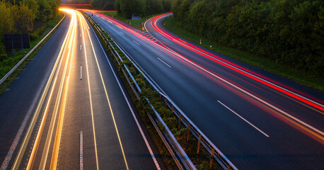 German motorway A46 at dusk at junction „Iserlohn-Zentrum“ in Sauerland. Colorful diagonal light traces on four lanes at evening twilight. Fast cars with yellow headlights and red lights in motion.