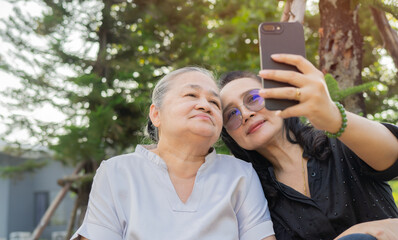 Cheeful adult daughter and mom sitting on bench and taking selfie in the park.