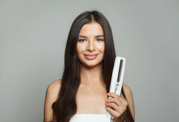 Nice young healthy brunette woman holding hair iron and straightening her long smooth shiny hair on white background. Haircare, hairstyle, hairdressing and hair styling concept