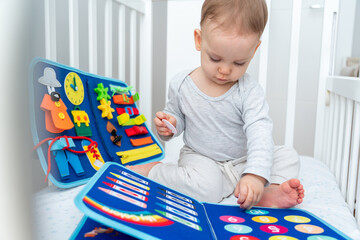 Little baby girl playing with busy book while sitting in crib. Concept of quiet books and modern educational toys - obrazy, fototapety, plakaty