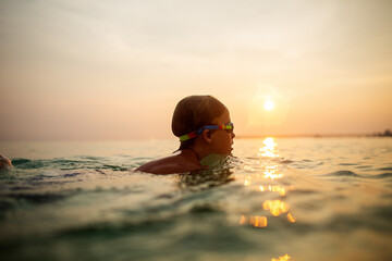 Calm child with goggles swimming at ocean sunset