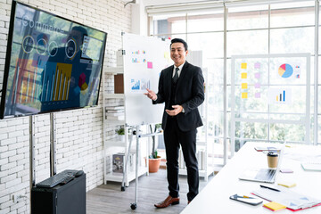 A cheerful and confident Asian businessman stands, presenting bar charts data from the projector screen to his office colleagues. Asian businessman leader role at the meeting.