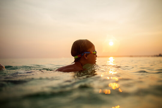 Calm child with goggles swimming at ocean sunset