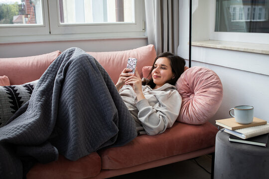 Young woman lying on coach and cozy plaid and scrolling on the phone