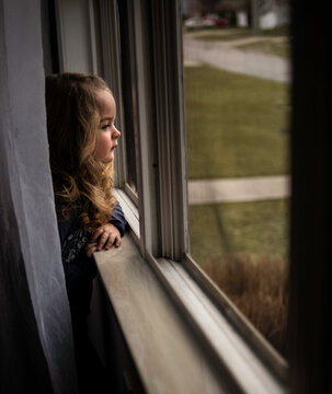 Beautiful little girl with blond curls looking out window in summer