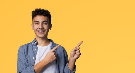 Excited happy black curly haired man in braces, wear denim shirt advertise show peek pointing area for sale slogan text, isolated over yellow background. Dental care ad advertisement concept. - obrazy, fototapety, plakaty