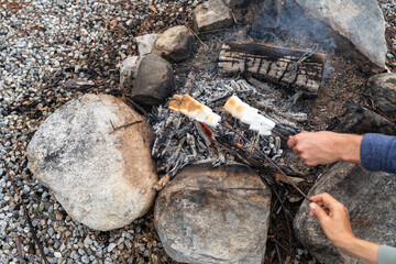 Two hands hold marshmallow sticks over the coals of a campfire. Cooking marshmallow meal on campfire for dessert for dinner. Travel food. Grill on the go. - obrazy, fototapety, plakaty