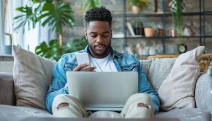 A man with a beard sits on a couch using a laptop and cell phone for leisure