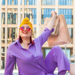 woman with craft shopping bags in the city. The concept of fashionable Consumerism and ecology. An adult Asian woman in a purple suit with red and yellow accents enjoys shopping . a sunny day.