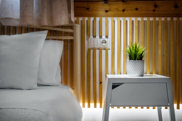 Captivating bedroom detail featuring part of bed adorned with crisp white bed linen, complemented by white bedside table adorned with green plant. A warm wood wall serves as a rustic backdrop