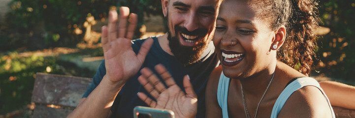 Happy smiling interracial couple taking selfie on phone while sitting on park bench, Panorama. - obrazy, fototapety, plakaty