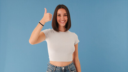 Smiling beautiful young brunette woman dressed in white top looking at camera showing thumbs up...