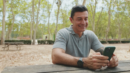Fototapeta premium Middle-aged man wearing gray T-shirt uses mobile phone while sitting on park bench
