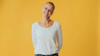Young hairless woman crossing her arms and looking at camera with smile, advertising space, isolated on yellow background in studio - obrazy, fototapety, plakaty
