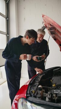 Vertical video of experienced car mechanic servicing car engine looking under hood with male trainee drinking hot drink - shot in slow motion