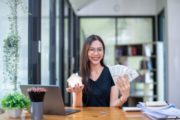 Smiling woman displaying a model house and a fan of cash, representing successful investment or...