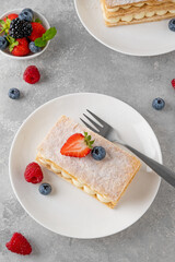 Traditional French dessert millefeuille with vanilla cream and fresh berries on a white plate on a gray concrete background.