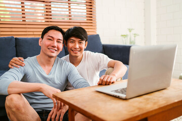 Happy millennial Asian gay couple sitting on a sofa while watching content on a laptop together in the living room at home. LGBT multi-relationship. Gay couple or same-sex marriage living together.