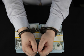Top view of handcuffed male hands lying on stacks of hundred dollar bills. On a black background,...