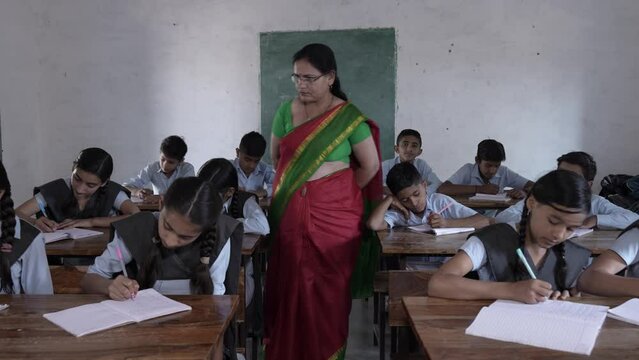 Female Indian teacher walks between desks at school during Exams, Elementary students boys and girls studying at classroom, Education in india.